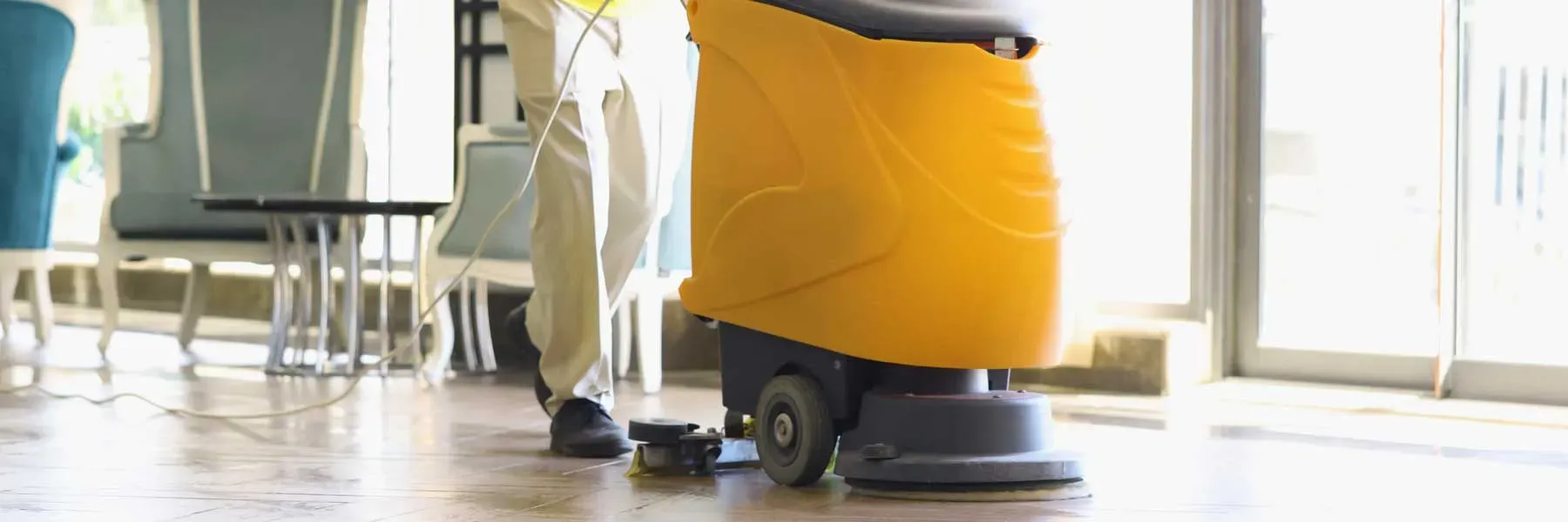 Cleaner operating a yellow floor scrubber in a commercial space near a seating area